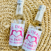 Two small clear glass bottles with white labels featuring pink text and a pink heart design, placed on a woven beige straw surface. The labels read 'Til Death Us Party Kelly's Bachelorette' with 'EST.
