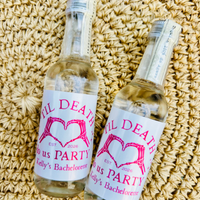 Two small clear glass bottles with white labels featuring pink text and a pink heart design, placed on a woven beige straw surface. The labels read 'Til Death Us Party Kelly's Bachelorette' with 'EST.