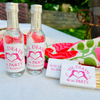 Two mini liquor bottles are featured beside matching matchboxes that feature a design with text reading 'Til Death Do Us Party' and a heart design, arranged on a white tablecloth outdoors with a colorful kitchen towel in the background