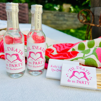 Two mini liquor bottles are featured beside matching matchboxes that feature a design with text reading 'Til Death Do Us Party' and a heart design, arranged on a white tablecloth outdoors with a colorful kitchen towel in the background