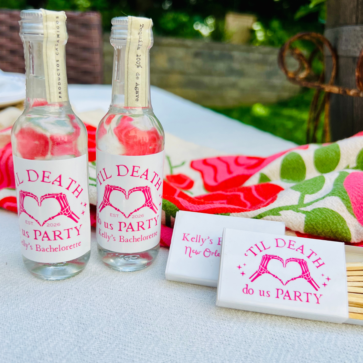 Two mini liquor bottles are featured beside matching matchboxes that feature a design with text reading 'Til Death Do Us Party' and a heart design, arranged on a white tablecloth outdoors with a colorful kitchen towel in the background