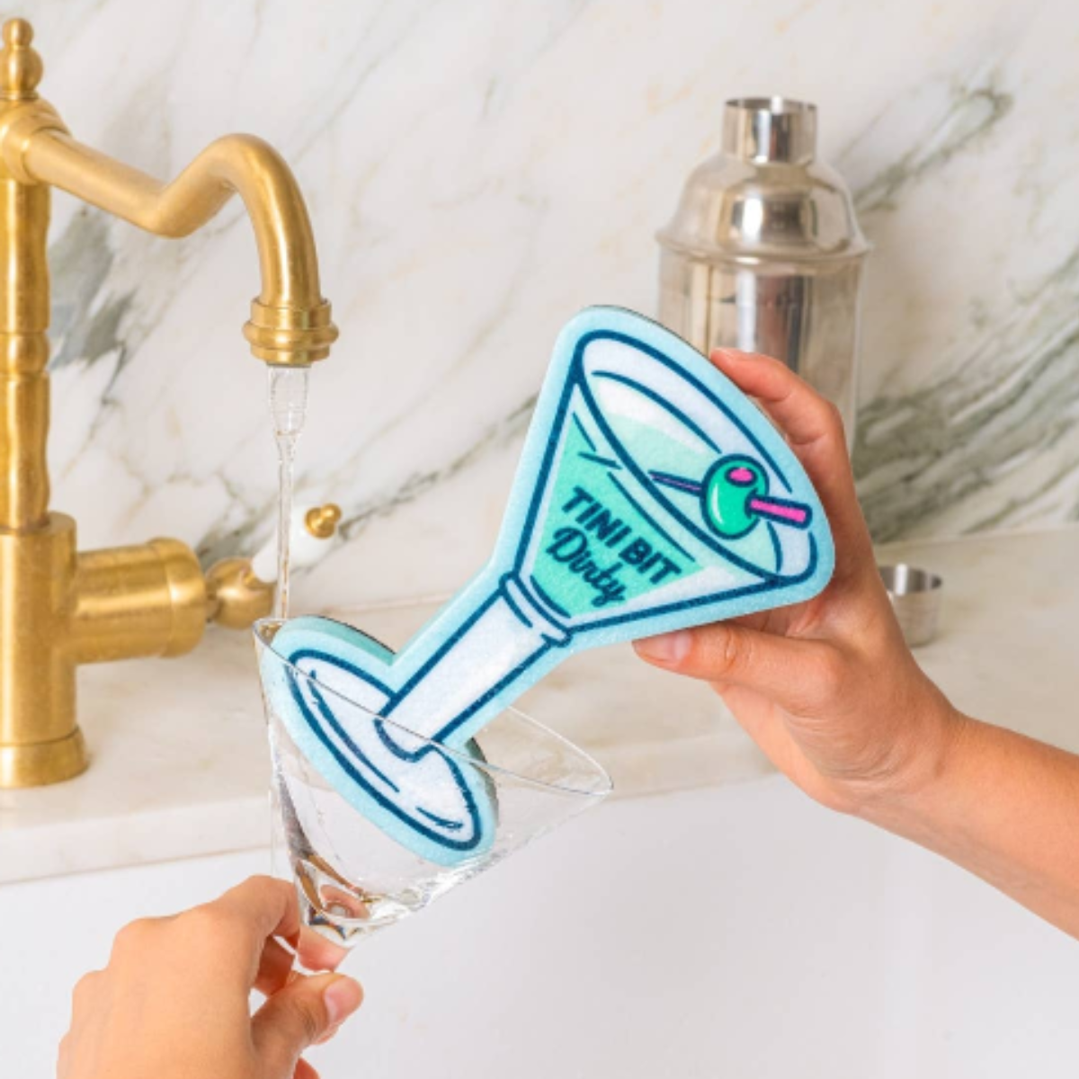 A person holds a cocktail-shaped sponge over a glass, with water running from a gold faucet in the background. The sponge is light blue with dark blue outlines and reads 'TINI BIT Dandy' with a small green olive detail.
