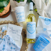 White wine bottle with a customized wine label featuring text that reads "Up to Snow Good" is shown on a table beside matching white and blue party cups, party favor bag, and napkins.