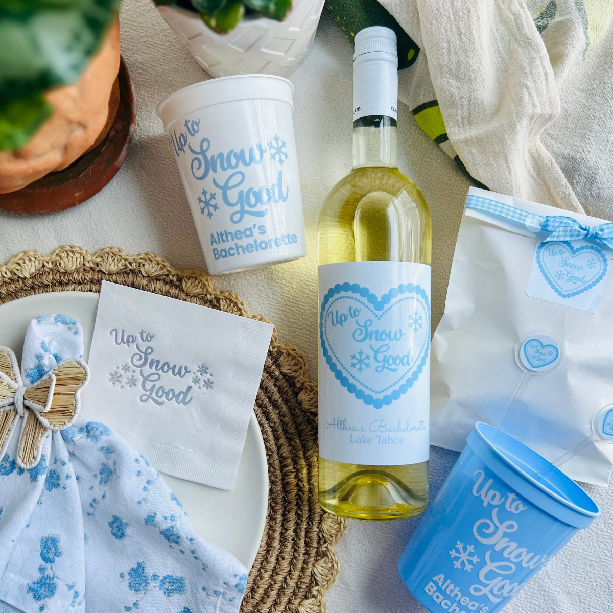 White wine bottle with a customized wine label featuring text that reads "Up to Snow Good" is shown on a table beside matching white and blue party cups, party favor bag, and napkins.