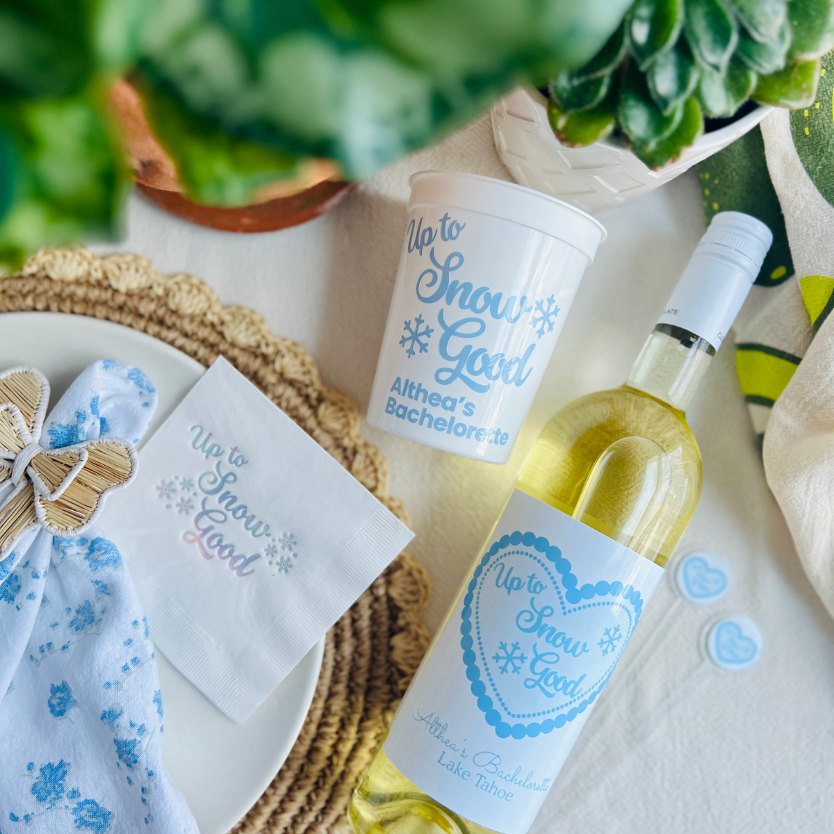 Bottle of white wine with 'Up to Snow Good' label, matching customized stadium bachelorette cups, and drink stirrers.