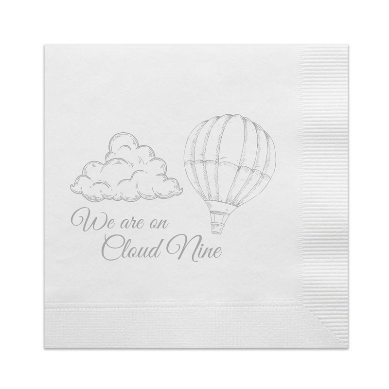White paper cocktail napkin with a silver foil stamped design featuring script that reads "We Are On Cloud Nine." The script appear below a fluffy cloud and a floating hot air balloon. A perfect napkin for a gender reveal party or a "On Cloud Nine" baby shower. 