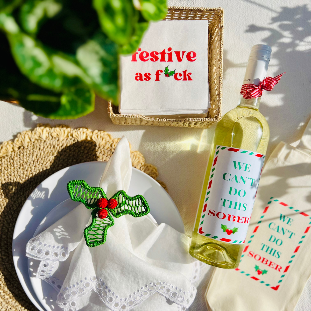 Overhead shot of a Christmas table decorated with "Festive as f*ck" white cocktail napkins and a wine bottle and matching wine bag with funny text that reads "We Can't Do This Sober." The canvas wine bag is a perfect way to wrap a bottle of wine for the holidays.