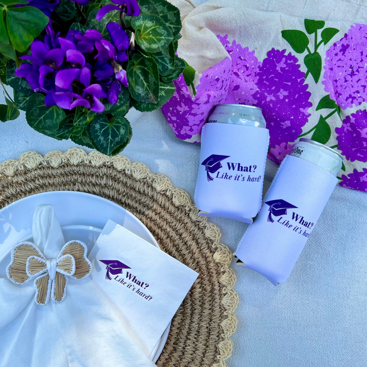 Two white can coolers with a matching design that features a graduation cap and text that reads "What? Like it's hard"" in purple  on a table with purple flowers and a woven placemat. There is a matching napkin with the same design on a dinner plate. 