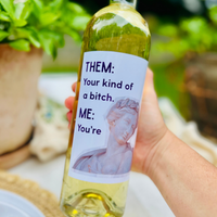 Hand holds a bottle of white wine in an outdoor setting. The wine bottle has a hilarious wine label that reads: "Them: Your kind of a bitch. Me: You're."