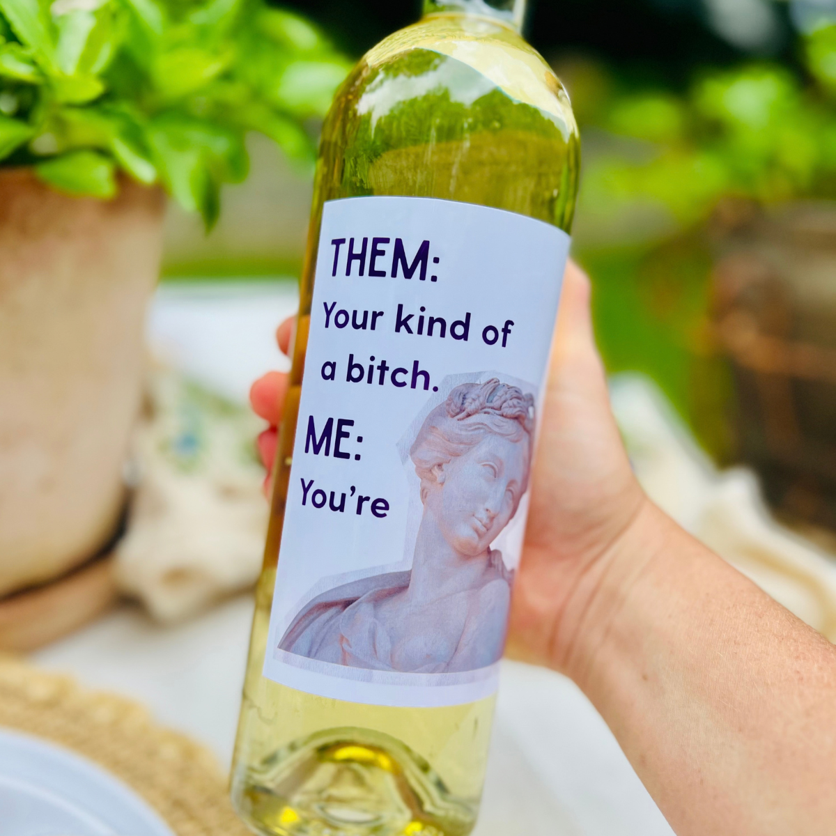 Hand holds a bottle of white wine in an outdoor setting. The wine bottle has a hilarious wine label that reads: "Them: Your kind of a bitch. Me: You're."